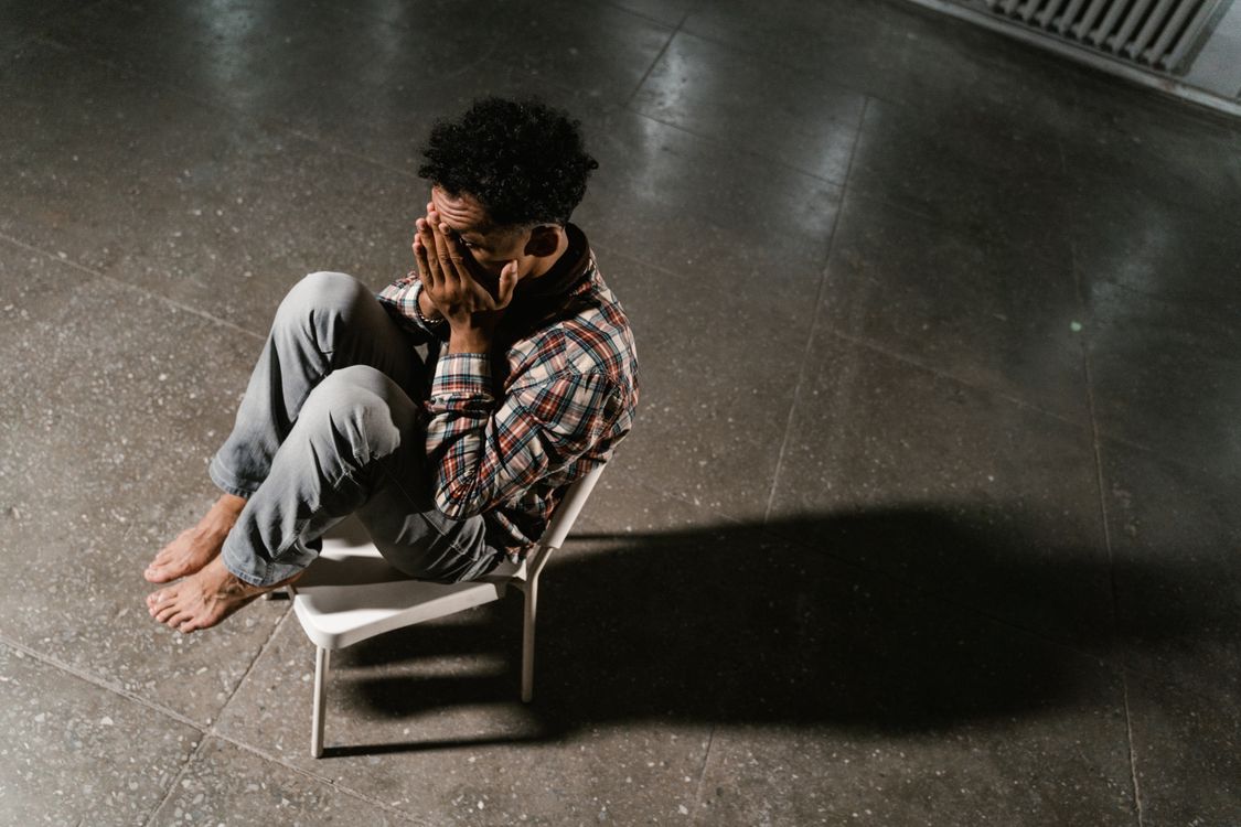A man sitting alone in a dim room, head down, showing signs of low confidence and stress